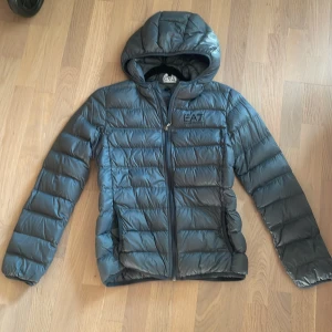 Mörkgrå dunjacka EA7 Emporio Armani - Snygg mörkgrå dunjacka från EA7 Emporio Armani med huva och dragkedja framtill. Jackan har ett stilrent, quiltat mönster och logga på bröstet. Perfekt för kyliga dagar och ger en clean look.