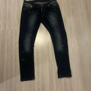Mörkblå jeans från Dsquared2 - Snygga mörkblå jeans från Dsquared2 med coola slitningar och vita färgstänk. Klassisk femficksmodell med metallogo bak och kontrastsömmar. Jeansen har en rak passform och är tillverkade i mjukt denimtyg för en bekväm känsla.