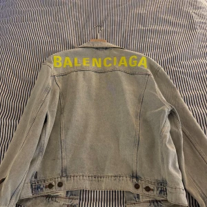 Balenciaga jeansjacka med gul logga - Säljer en ljusblå jeansjacka från Balenciaga med stor gul logga broderad på ryggen. Klassisk modell med knappar framtill, två bröstfickor med lock och två sidofickor. Jackan har en loose fit och är tillverkad i denim, storleken är 38 men sitter mer som en oversized M (se bild) är 188cm 86kg jackan är köpt på Myth o Theresa, kvitto finns!