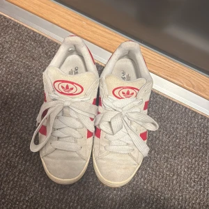Adidas sneakers med röda ränder - Säljer ett par klassiska Adidas sneakers i vitt med röda ränder och Adidas-logga på plösen. Skorna har snörning, gummisula i brunt och är tillverkade i skinn och syntet. Perfekta för dig som gillar retrostil och streetwear.