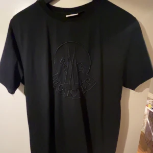 Svart Moncler t-shirt med broderad logga - Säljer en svart t-shirt från Moncler med stor broderad logga på bröstet och klassisk Moncler-patch på ärmen. T-shirten har rund hals och korta ärmar, perfekt för en clean och stilren look. Materialet känns mjukt och skönt mot huden. Köpt i marbella föra sommaren 