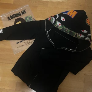 Bare Hoodie - Svart hoodie från BAPE med ikonisk shark-hood, camo-detaljer och broderade patchar på huvan. Dragkedja framtill, ribbade muddar och tryckt logga på ärmen. Perfekt för dig som vill sticka ut med streetwear-vibe.