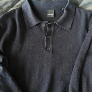 Mörkblå stickad pikétröja Only & Sons - Snygg mörkblå stickad pikétröja från Only & Sons med klassisk krage och tre knappar framtill. Tröjan har lång ärm och är perfekt för dig som gillar en stilren och enkel look. Passar till både jeans och chinos.