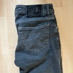 Mörkgrå jeans från Jacob Cohen - Snygga mörkgrå jeans från Jacob Cohen med raka ben och klassisk femficksdesign. Byxorna har silverfärgade knappar, broderad logga på fickan och svart patch bak i midjan. Tillverkade i mjukt bomullstyg med exklusiv känsla. Perfekta för dig som gillar stilrena och tidlösa jeans.