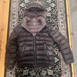 Brun dunjacka från Colmar - Snygg mörkgrå dunjacka från Colmar med huva och två snedställda fickor med dragkedja. Jackan har quiltad design och grov dragkedja framtill, perfekt för kalla dagar. Stilsäker look med diskret logga på ärmen och hög krage. Mycket fint skick endast en lagning på insidan av innerfickan som ej syns vid användning och mycket små defekter på ena armen. Hör av dig vi intresse😊