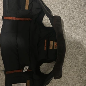 Svart dunväst från Parajumpers - Svart dunväst från Parajumpers med hög krage fodrad i brun fuskpäls, dragkedja och knappar framtill samt praktiska fickor med dragkedja. Västen har klassisk Parajumpers-logga bak i nacken och orange detaljer på insidan. Storlek xs men passar S 