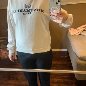 Vit sweatshirt Montmartre Paris - Vit sweatshirt från Montmartre Paris med svart tryck framtill. Tröjan har rund hals, långa ärmar och en avslappnad passform. Perfekt för dig som gillar enkel och stilren streetwear.