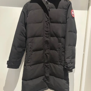 Svart jacka - Säljer en svart, lång jacka från C*nada g**se med klassisk patch på ärmen och fluffig päls runt huvan. Jackan är dock köpt second hand så kan inte garantera att den är äkta därav priset, men enligt deras kundtjänst kundtjänst verkar allt stämma. Jackan har dragkedja, knappar och två stora fickor framtill. Perfekt för kalla vinterdagar. Har en reva på insidan i den högra armhålan men inget som syns. Använd men i fint skick och väldigt varm. Passar xs-s