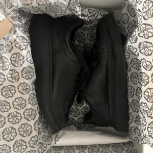 Svarta sneakers Alexander McQueen - Snygga helsvarta sneakers från Alexander McQueen i skinn med rund tå och bred platt sula. Klassisk modell med svarta skosnören och diskret guldfärgad logga på plösen och hälen. Perfekta för dig som gillar stilrena och exklusiva sneakers.