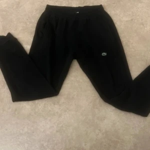 Svarta mjukisbyxor från Lacoste - Svarta mjukisbyxor från Lacoste med regular fit och klassisk krokodillogga på benet. Byxorna har resår i midjan och är tillverkade i mjukt material som passar perfekt för chill eller träning. Snygg och enkel design som funkar till det mesta.