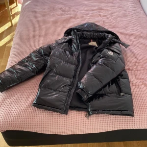 Svart Moncler pufferjacka med huva - Snygg svart pufferjacka från Moncler med glansig finish och klassisk logga på ärmen. Jackan har huva, dragkedja framtill och två sidofickor med dragkedja. Perfekt för kalla dagar när du vill hålla stilen och värmen.