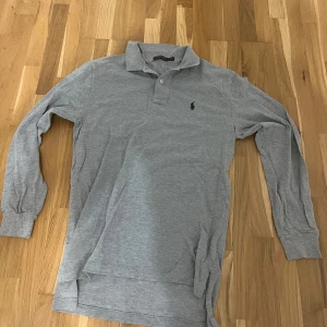 Grå pikétröja från Polo Ralph Lauren - Klassisk grå långärmad pikétröja från Polo Ralph Lauren med broderad marinblå logga på bröstet. Tröjan har krage, knäppning med två knappar och är tillverkad i mjuk bomull. Perfekt för en clean och stilren look.