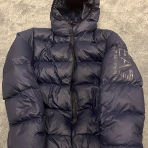 EA7 Emporio Armani vinterjacka - Snygg mörkblå pufferjacka från EA7 Emporio Armani med stor logga på ärmen. Jackan har huva, dragkedja framtill och quiltad design. Perfekt för kalla dagar och ger en clean streetstyle-look.