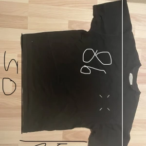 Maison Margiela svart t-shirt med tryck - Svart t-shirt från Maison Margiela med ikoniskt siffertryck och stor 6:a på ryggen. Klassisk passform, rund hals och korta ärmar. Tillverkad i mjuk bomull, perfekt för dig som gillar streetwear och designerplagg.