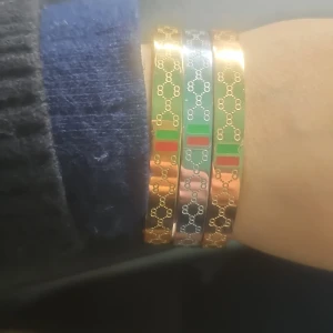  armband i guld och silver - Tre stilrena armband i metall med mönster. Två armband är guldfärgade och ett är silverfärgat, alla med gröna och röda detaljer. Perfekt accessoar för dig som vill sticka ut med lyxig känsla och trendig design.