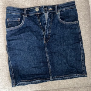 Mörkblå jeanskjol från Zara - Snygg mörkblå jeanskjol från Zara med klassisk femficksdesign, bälteshällor och dragkedja framtill. Kjolen har raka linjer och är i ett mjukt denimtyg som ger en avslappnad men stilren look. Perfekt att matcha med en enkel t-shirt eller hoodie.