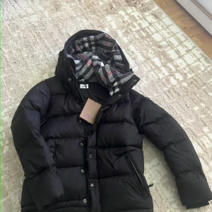 Svart dunjacka från Burberry - Svart pufferjacka från Burberry med avtagbara ärmar, vilket gör att den kan bäras som väst. Jackan har en huva fodrad med klassiskt rutigt tyg i grått, vitt och rött. Två dragkedjeförsedda fickor framtill och stängs med både dragkedja och knappar.