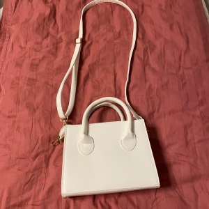 Vit axelväska med handtag - Stilren vit axelväska i syntetmaterial med både justerbar axelrem och två handtag. Väskan har en fyrkantig form och stängs med dragkedja i guld. 