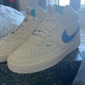 Nike Court Vision Mid vit/blå/grön - Nike Court Vision Mid sneakers i vitt skinn med blå swoosh och gröna detaljer på plös och häl. Klassisk perforerad tå, snörning och platt sula. Fräscha färgkombinationer och retro basketstil som sticker ut. Perfekt för dig som gillar sportig vibe och snygga detaljer. Inte använd svara testat dem o j då insett att dem inte passar mig så därför säljer jag dem sänker kanske vid snabb affär
