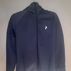 Peak Performance zipper  - Säljer en marinblå hoodie från Peak Performance med hel dragkedja och huva. Tröjan har vit logga på bröstet och texten 'PERFORMANCE' tryckt runt huvan. Perfekt för chill eller träning, med två fickor framtill.