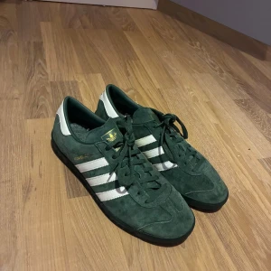 Adidas Hamburg gröna sneakers mocka - Säljer ett par Adidas Hamburg sneakers i mörkgrön mocka med vita detaljer och klassiska tre ränder på sidorna. Skorna har svart sula, snörning och guldfärgad Hamburg-text på sidan. Perfekta för dig som gillar retrostil och streetwear.
