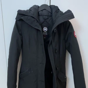 Svart dunjacka från Canada Goose 2XS - Säljer en svart dunjacka från Canada Goose i storlek 2XS. Jackan har huva, dragkedja och knappar framtill samt klassisk logga på ärmen. lång modell med praktiska fickor. Box och galgen finns kvar, helt ny då den bara var använd en gång förra vintern sen bytte jag jacka så bara legat hemma. Nypris 14000