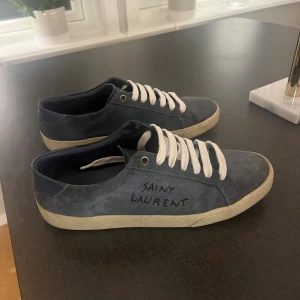 Saint Laurent blå sneakers mocka - Snygga blå sneakers från Saint Laurent i mocka med vit sula och vita snören. Klassisk låg modell med rund tå och diskret logga på sidan. Perfekta för dig som gillar stilrena och exklusiva skor.