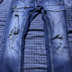 Blå dsqured2 jeans från Denim Park - Säljer ett par blå jeans från Denim Park med rejält slitna detaljer och hål framtill. Jeansen har färgstänk i gult och vitt för en extra edgy look. Klassisk femficksmodell med normal passform och raka ben. Perfekta för dig som gillar streetstyle.