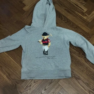 Grå hoodie Polo Bear Ralph Lauren - Grå hoodie från Ralph Lauren med ikoniska Polo Bear-trycket framtill. Klassisk huva, långa ärmar och mjukt material. Perfekt för dig som gillar streetstyle och vill ha en chill look med en touch av sportig vibe.