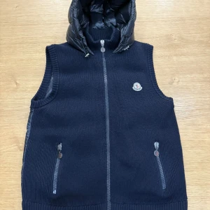 ÄKTA! Mörkblå Tricot dunväst från Moncler - Snygg mörkblå dunväst från Moncler med stickad utsida och glansig quiltad huva. Västen har dragkedja framtill, två fickor med dragkedja och Moncler-logga på bröstet. Perfekt för lager-på-lager och streetstyle. Använd men i ny skick  