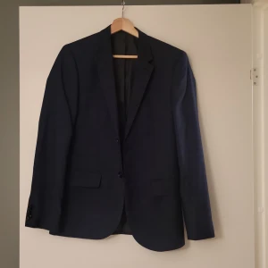 Mörkblå kavaj Skinny Fit från H&M - Säljer en mörkblå kavaj från H&M i modellen Skinny Fit. Kavajen har två knappar framtill, klassiska fickor med lock och smal passform. Perfekt för dig som gillar en stilren och modern look. Materialet är mjukt och lätt, vilket gör den bekväm att bära.