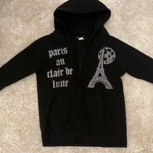 Svart hoodie med strass och Paris-tryck - Svart hoodie från Bling lucid  i storlek 12/13 år XL i barn storlek med dragkedja, huva och fickor. Framsidan har strassdetaljer med texten 'paris au clair de lune' och motiv av Eiffeltornet och månen. Perfekt statement-plagg för dig som gillar Paris-vibes och bling, säljer då den är för liten! 