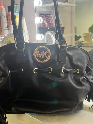 Svart handväska Michael Kors - Snygg svart handväska från Michael Kors med guldiga detaljer och MK-logga framtill. Väskan har två långa axelremmar och snörning med metallringar. Tillverkad i skinn och har en mjuk, rymlig form som gör den perfekt för att bära med sig allt man behöver.