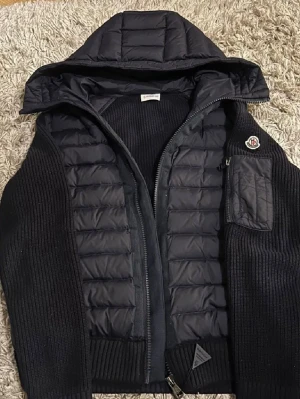 Svart pufferjacka från Moncler - Svart pufferjacka från Moncler med stickade ärmar och quiltad kropp. Jackan har huva, dragkedja framtill och ribbade muddar. Snygg Moncler-logga på ena ärmen och praktiska fickor med knappstängning. Perfekt för kyliga dagar och riktigt stilren.