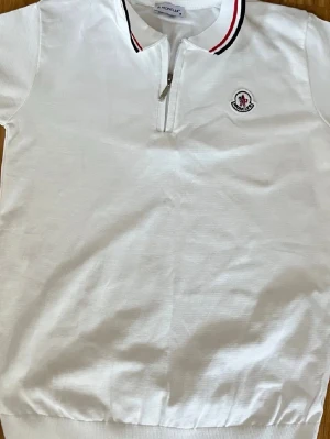 Vit Moncler polotopp med dragkedja - Säljer en vit polotopp från Moncler med korta ärmar och dragkedja framtill. Kragen har snygga ränder i rött och svart, och på bröstet finns Moncler-loggan broderad. Toppen är gjord i mjuk bomull och har en klassisk, sportig look. 