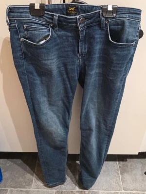 Blå jeans från Lee, straight fit - Säljer ett par klassiska blå jeans från Lee med straight fit och fem fickor. Jeansen har normal passform och är tillverkade i slitstarkt denimtyg. Snygga detaljer med Lee-logga på bakfickan och metallknapp framtill. Perfekta för dig som gillar tidlös stil.