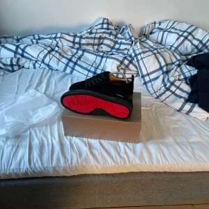louboutin skor - Säljer ett par svarta sneakers från Christian Louboutin med ikonisk röd sula och snörning. Skorna har en stilren och modern design med rund tå och platt sula. Perfekta för dig som vill sticka ut med exklusiva detaljer.