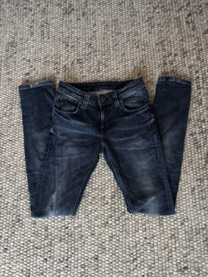 Mörkblå jeans från Nudie Jeans - Snygga mörkblå jeans från Nudie Jeans med klassisk femficksdesign och bruna detaljer på bakfickan. Jeansen har en slim passform och är tillverkade i slitstarkt denim med coola tvättade effekter. Perfekt för dig som gillar stilrena och moderna jeans.