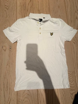 Vit piké från Lyle & Scott - Klassisk vit pikétröja från Lyle & Scott med korta ärmar, krage och två knappar framtill. Ikonisk gul logga på bröstet. Tillverkad i mjuk bomull som känns skön mot huden. Perfekt till jeans eller shorts för en clean look.