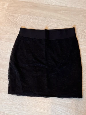 Svart spetskjol med elastisk midja - Säljer en svart kortkjol med snygg spetsdetalj och bred elastisk midja. Kjolen har en rak och tight passform som ger en cool och stilren look. Perfekt att matcha med en enkel topp och boots för en trendig outfit.