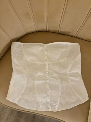 Vit korsett-topp/ tubtopp med knappar - Säljer en vit korsett-topp i glansigt material med snygga paneler och smockade partier. Toppen är axelbandslös och har dekorativa knappar framtill samt dragkedja i sidan för enkel på- och avtagning. Perfekt för dig som gillar stilrena och trendiga plagg.