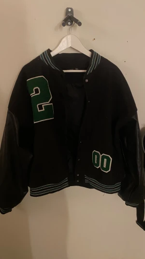 Svart bomberjacka från Boohoo - Svart bomberjacka från Boohoo med gröna och vita anlag i form av siffror på bröstet, tryckknappar framtill och tyvärr fejk fickor. Ärmarna är i skinn och kroppen i tyg. Perfekt för streetstil.💗 säljs pga att den inte används