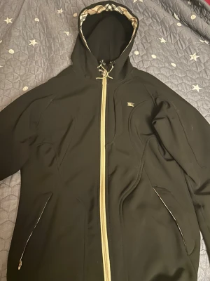 Svart Burberry vindjacka XL/sommarjacka - Svart vindjacka från Burberry i storlek XL med klassiskt rutigt mönster på insidan av huvan och längs dragkedjan. Jackan har snörning vid huvan, guldfärgad logga på bröstet och praktiska fickor med dragkedja. Perfekt för dig som gillar stilrena och lyxiga detaljer. Kan även användas som storlek S.