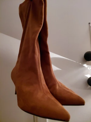 Bruna mockastövletter med spetsig tå - Säljer ett par bruna stövletter i mocka med spetsig tå och smal klack. Skorna har en elegant och stilren design som passar perfekt till höst- och vinterlooken. De är höga och sitter snyggt på benet, vilket ger en trendig siluett. Storlek 39.