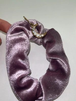 Ljuslila scrunchie med månberlock - Säljer en scrunchie i glansigt ljuslila tyg med en guldfärgad månberlock dekorerad med små stenar. Perfekt accessoar för håret eller handleden, ger en söt och trendig touch till din look.💜💜💜