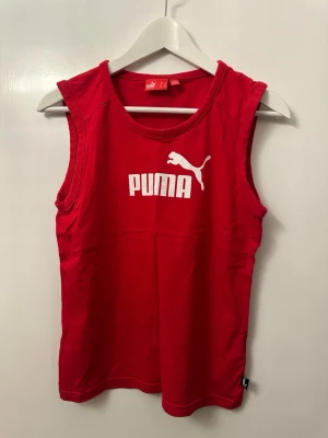 Röd linnetopp från Puma - Säljer en röd linnetopp från Puma med vitt tryck och logga framtill. Toppen är ärmlös med rund halsringning och tillverkad i mjuk bomull, perfekt för träning eller chill dagar.