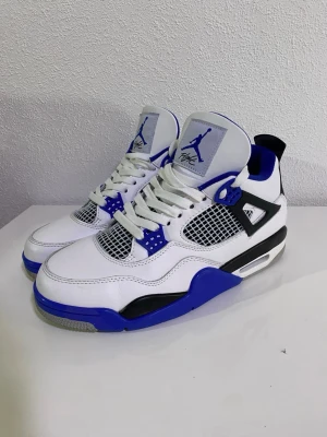 Nike Air Jordan 4 Retro blå/vit - Nike Air Jordan 4 Retro sneakers i vit och blå med svarta detaljer. Skorna har meshpaneler, snörning och Jumpman-logga på plös och häl. Snygg kontrast mellan blå och svart på sula och häl. Tillverkade i skinn och syntetmaterial, med platt sula och rund tå.