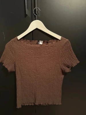 Brun ribbad topp med kort ärm - Säljer en brun ribbad topp från SHEIN med korta ärmar och stretchigt, smockat tyg. Toppen har en croppad passform och vågig kant vid halsringning, ärmslut och nederkant. Perfekt till jeans eller kjol för en avslappnad look.