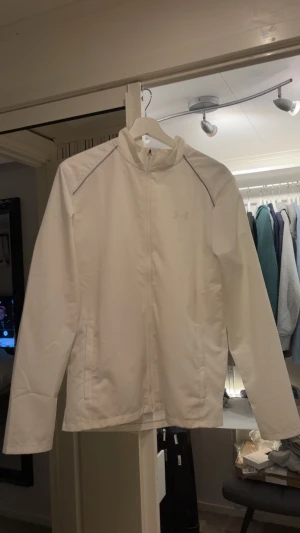 Vit vindjacka från Under Armour - Snygg vit vindjacka från Under Armour med diskret logga på bröstet och stor logga på ryggen. Jackan har dragkedja framtill, två fickor och tunna reflekterande detaljer vid axlarna. Perfekt för dig som gillar sportig stil och vill ha något lätt och fräscht.