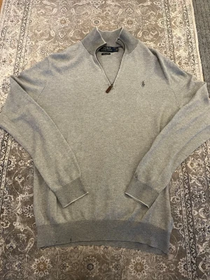 Grå stickad tröja Polo Ralph Lauren - Snygg grå stickad tröja från Polo Ralph Lauren med half zip och diskret logga på bröstet. Tröjan har långärmade ribbade muddar och en klassisk krage. Perfekt för dig som gillar stilrena och tidlösa plagg.